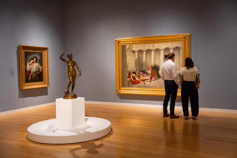 Two people viewing a large classical painting in a museum room with wooden floors and muted blue walls, next to a bronze statue on a white pedestal and another framed painting.
